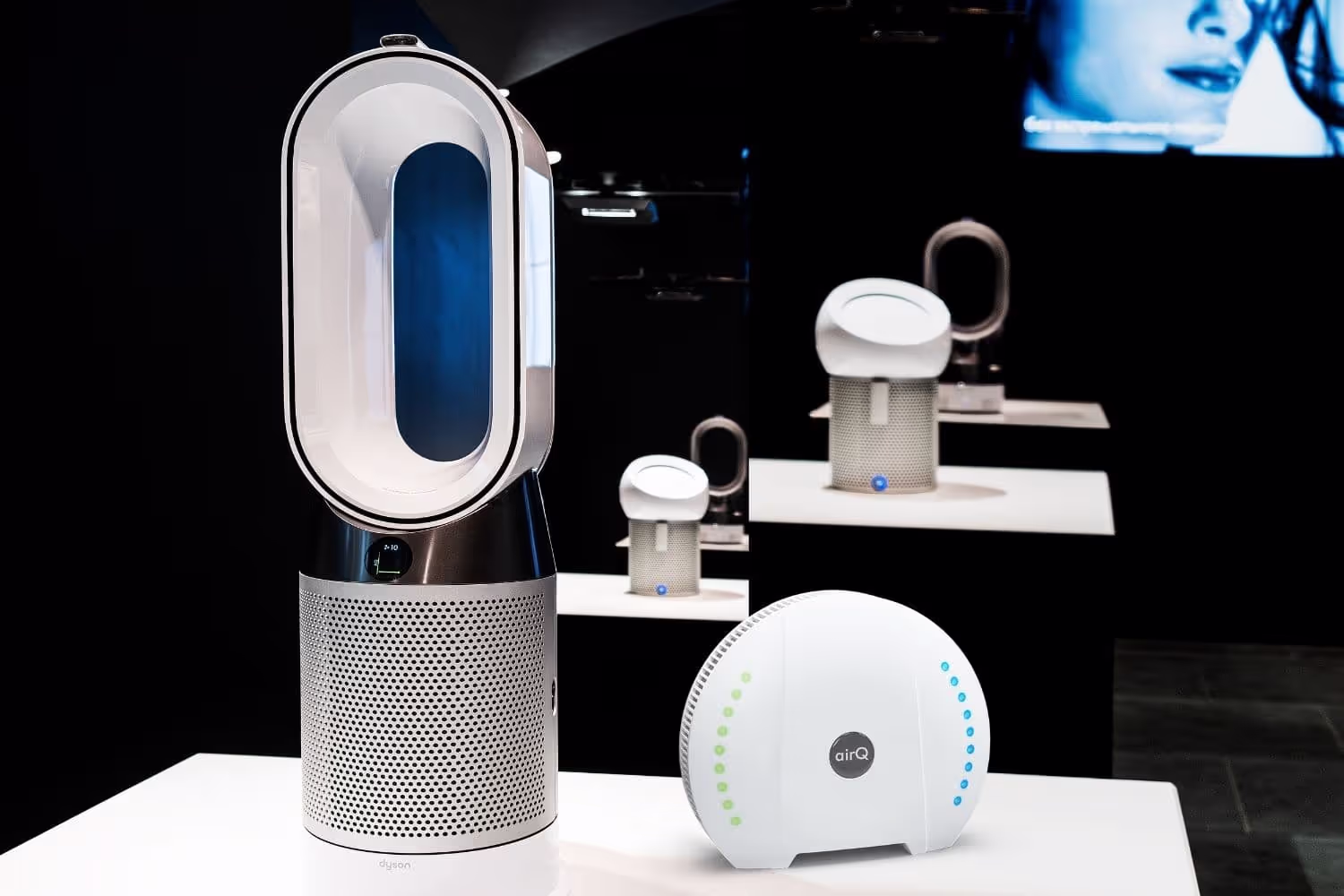 Luftreiniger Dyson und Luftmessgerät air-Q auf einem weißen Tisch stehend vor dunklem Hintergrund.