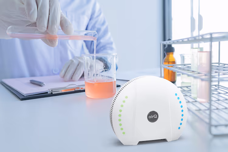 Labortisch mit Reagenzgläsern und dem air-Q mit einem VOC-PID-Sensor im Vordergrund. Im Hintergrund befüllt ein Laborassistent eine Flüssigkeit in einen Messbecher.