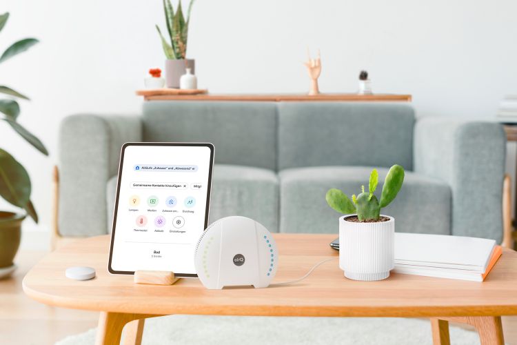Graues ofa im Hintergrund, im Vordergrund steht das Luftmessgert air-Q neben einem Tablet auf dem die air-Q App geöffnet ist auf einem Holztisch