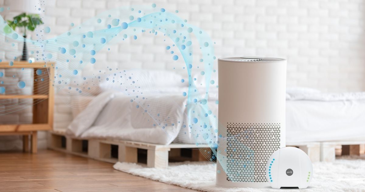 Helles Schlafzimmer mit Palettenbett und Nachttisch im HIntergrund, im Vordergrund steht ein Lufteiniger aus dem blaue Kugeln als Grafik strömen und das Luftmessgerät air-Q