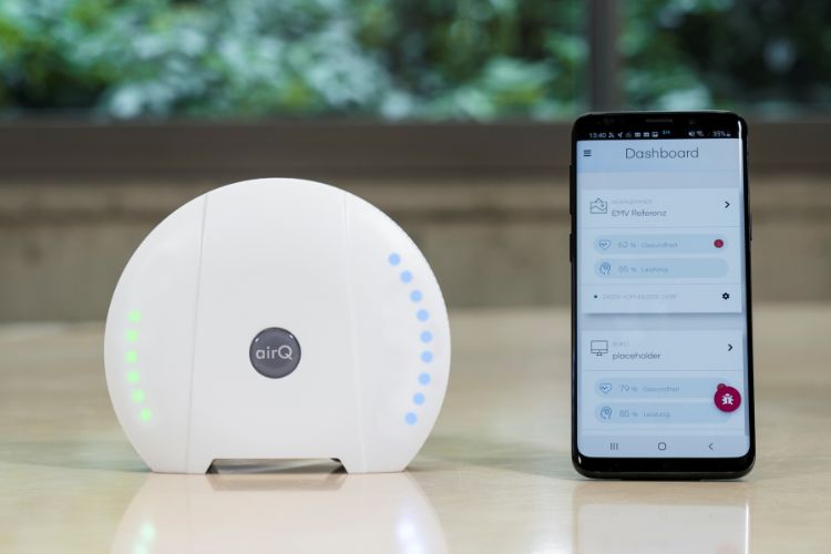 Luftgütesensor air-Q neben einem Smartphone mit geöffneter Dashboard-App, die Luftqualitätsdaten anzeigt, auf einem Tisch vor einem Fenster mit Blick ins Grüne."