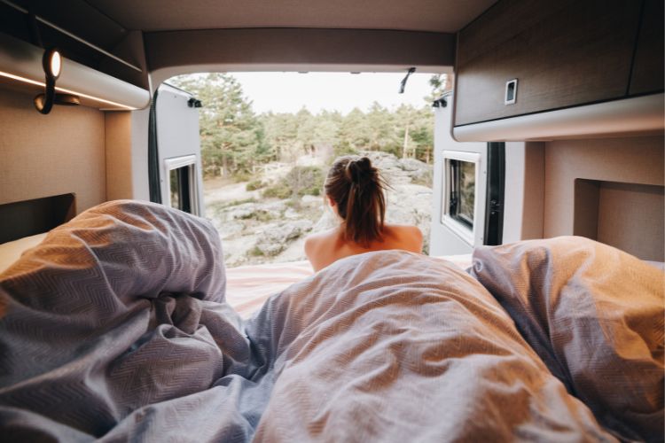 Campervan mit geöffneter Rücktür und einem Schlafplatz, in dem eine brünette Frau liegt und nach draußen schaut.