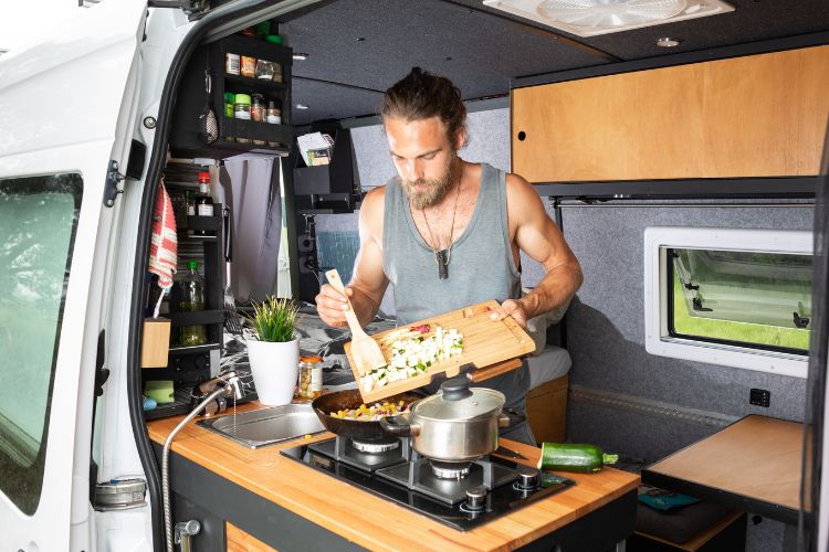 Geöffneter Campervan mit Kochzeile, an der ein junger Mann geschnittenes Gemüse in eine Pfanne gibt.
