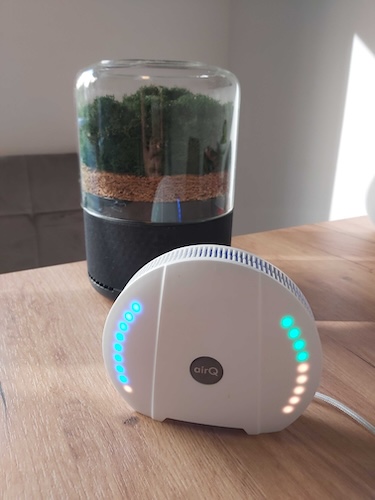 Luftmessgerät air-Q mit leuchtender LED-Anzeige und Luftreiniger Briiv im Hintergrund
