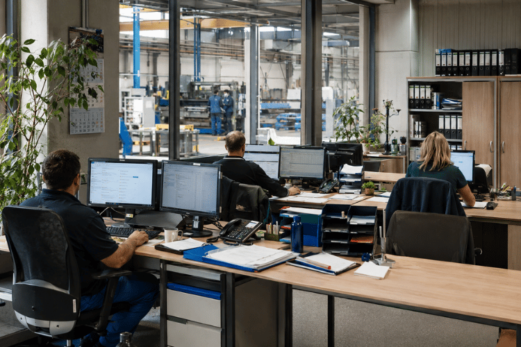 Varios puestos de trabajo de oficina en una empresa industrial moderna. Grandes ventanales permiten ver la nave de producción situada detrás, con máquinas y empleados trabajando en la fabricación.
